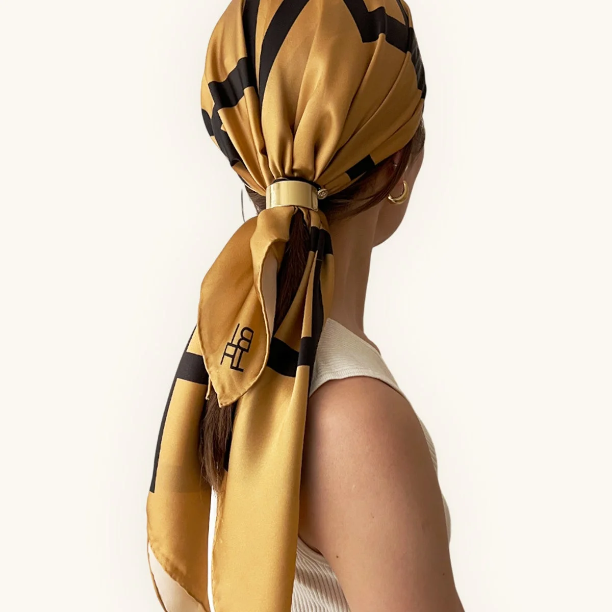 後ろ姿の女性が、ゴールドと黒の幾何学模様のスカーフを金色のリングで束ねて髪にアレンジしている様子