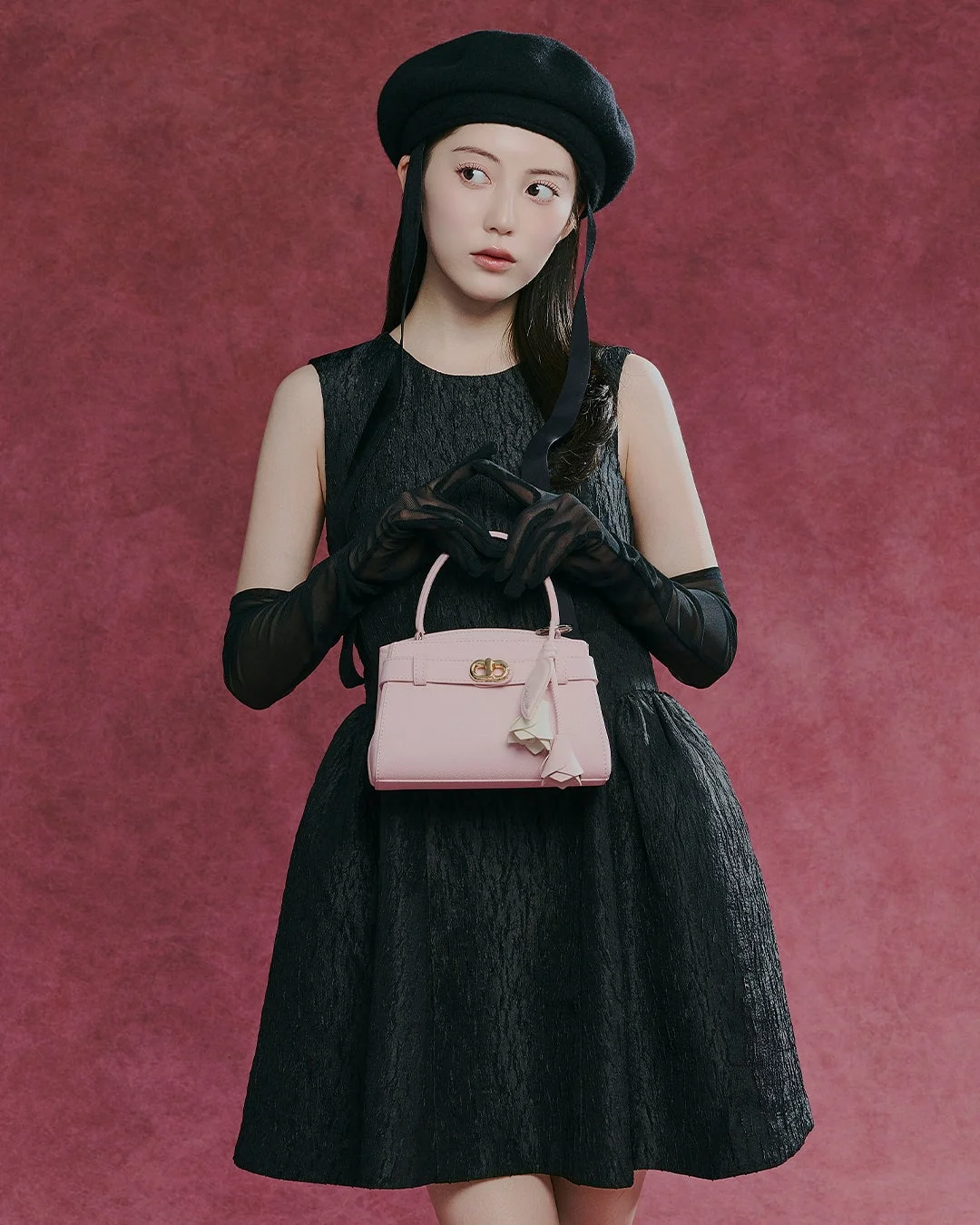 黒いベレー帽とドレスを着用し、透け感のある黒い手袋とピンクのハンドバッグを持った若い女性の全身ポートレート