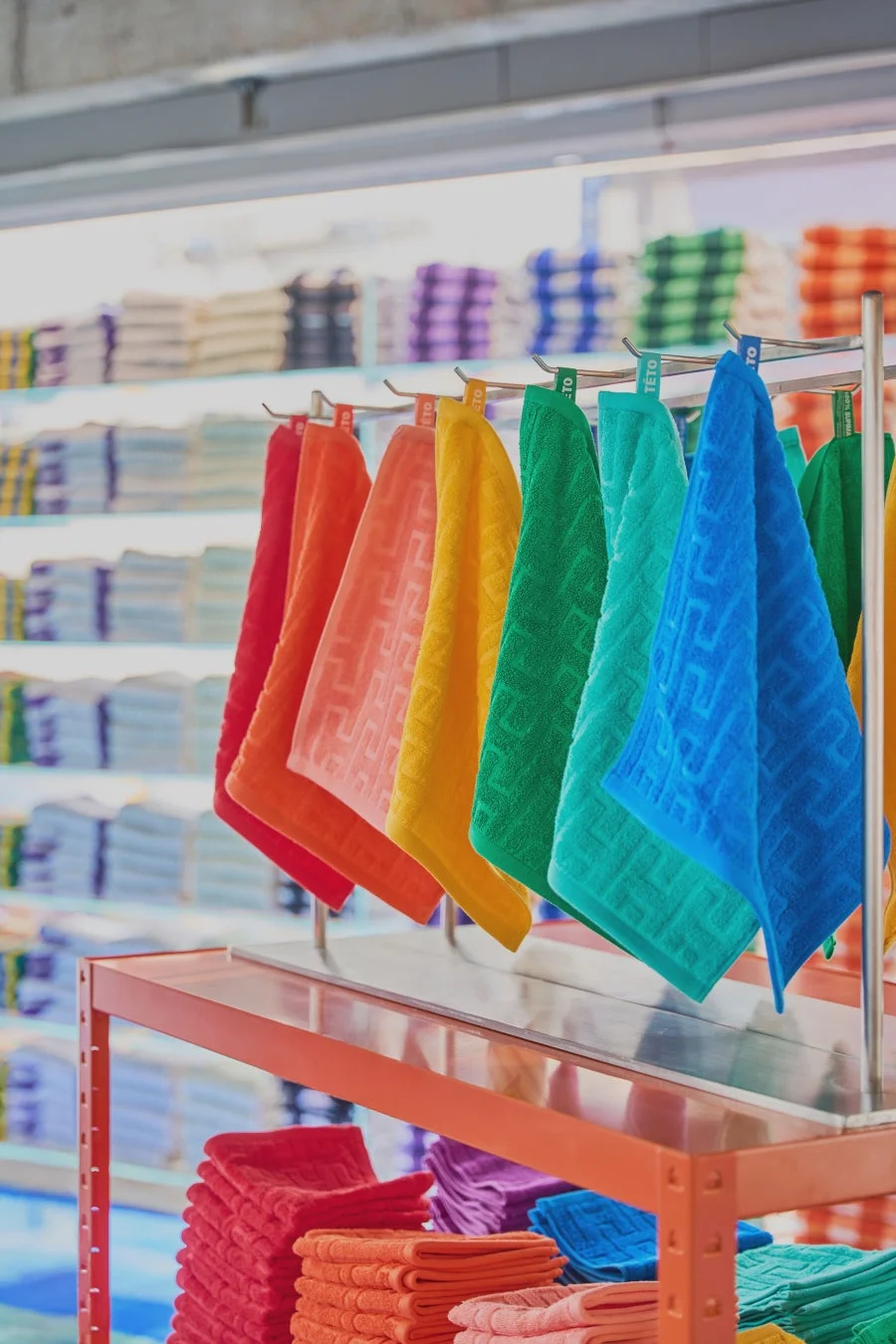 店舗の陳列棚に、赤、オレンジ、黄、緑、青など虹色のカラフルなタオルが並べられています。ハンガーに掛けられたものと積み重ねられたものがあり、清潔感と多様性を感じさせる商品ディスプレイです。