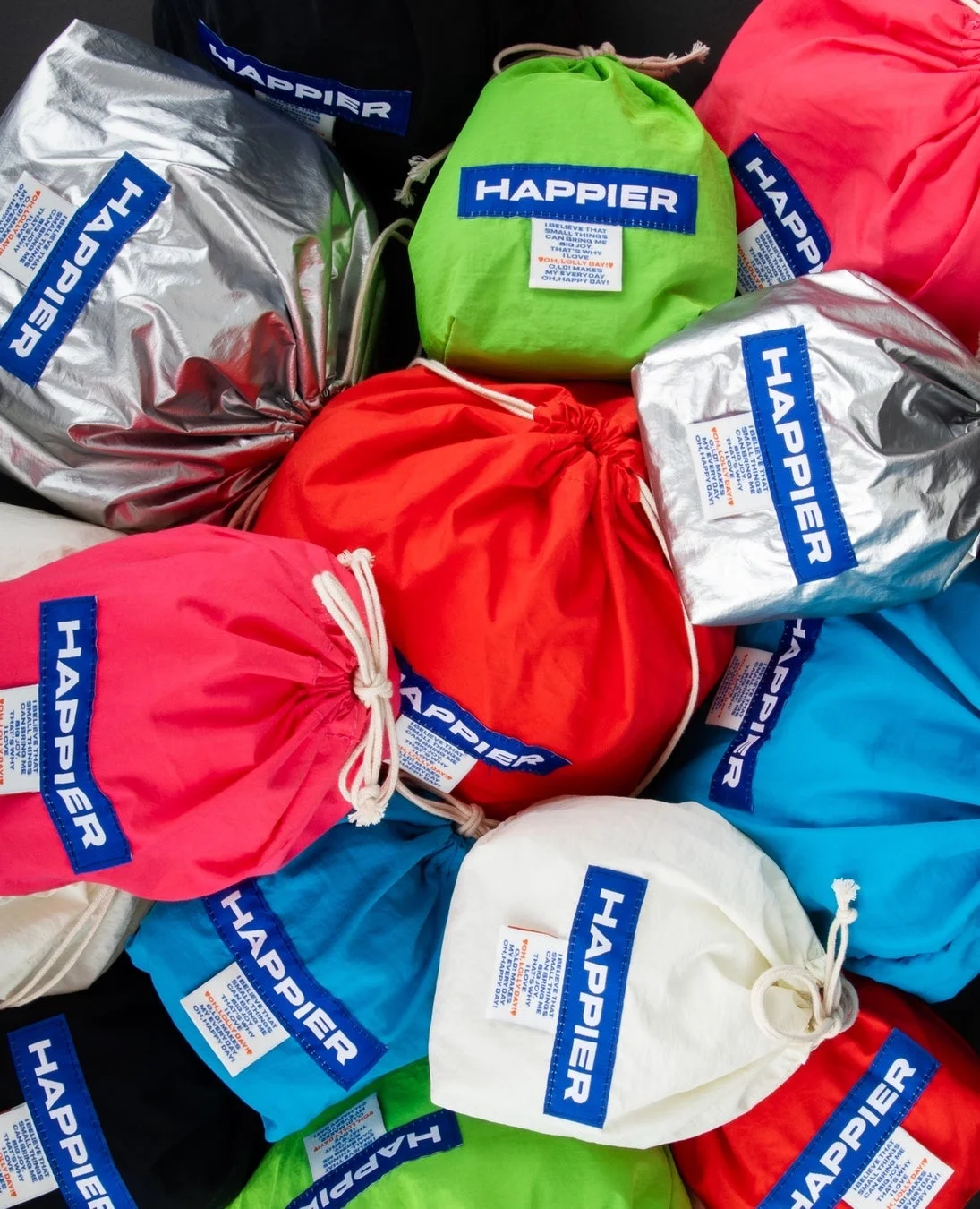 「HAPPIER」のロゴが入った、様々な色の巾着袋が山積みにされている様子。銀色、黄緑、赤、ピンク、水色、白など、鮮やかな色合いが特徴的で、明るく楽しい雰囲気を伝えています。