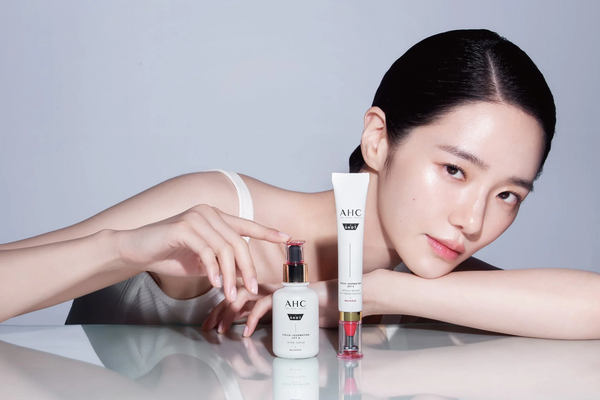 透明感のある肌の女性が、AHCブランドの美容液とアイクリームの2つのスキンケア製品と共にポーズをとっているスタジオ写真です。