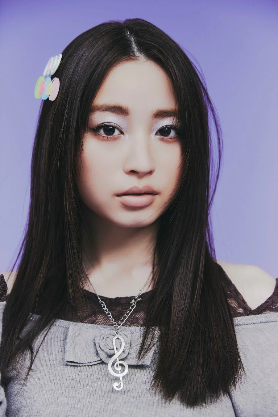 紫色の背景の前でポーズをとる、長い黒髪の若い女性のポートレート。グレーのトップスにト音記号のネックレスを着用し、カラフルなヘアクリップが特徴的。クールな表情が印象的です。