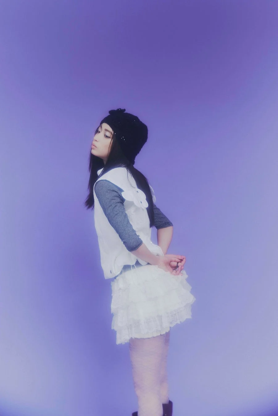 紫色のシンプルな背景の前で、黒いニット帽と白いフリルスカートを着用した女性が横向きに立っているポートレート。手を後ろで組んでおり、クールな表情が印象的です。