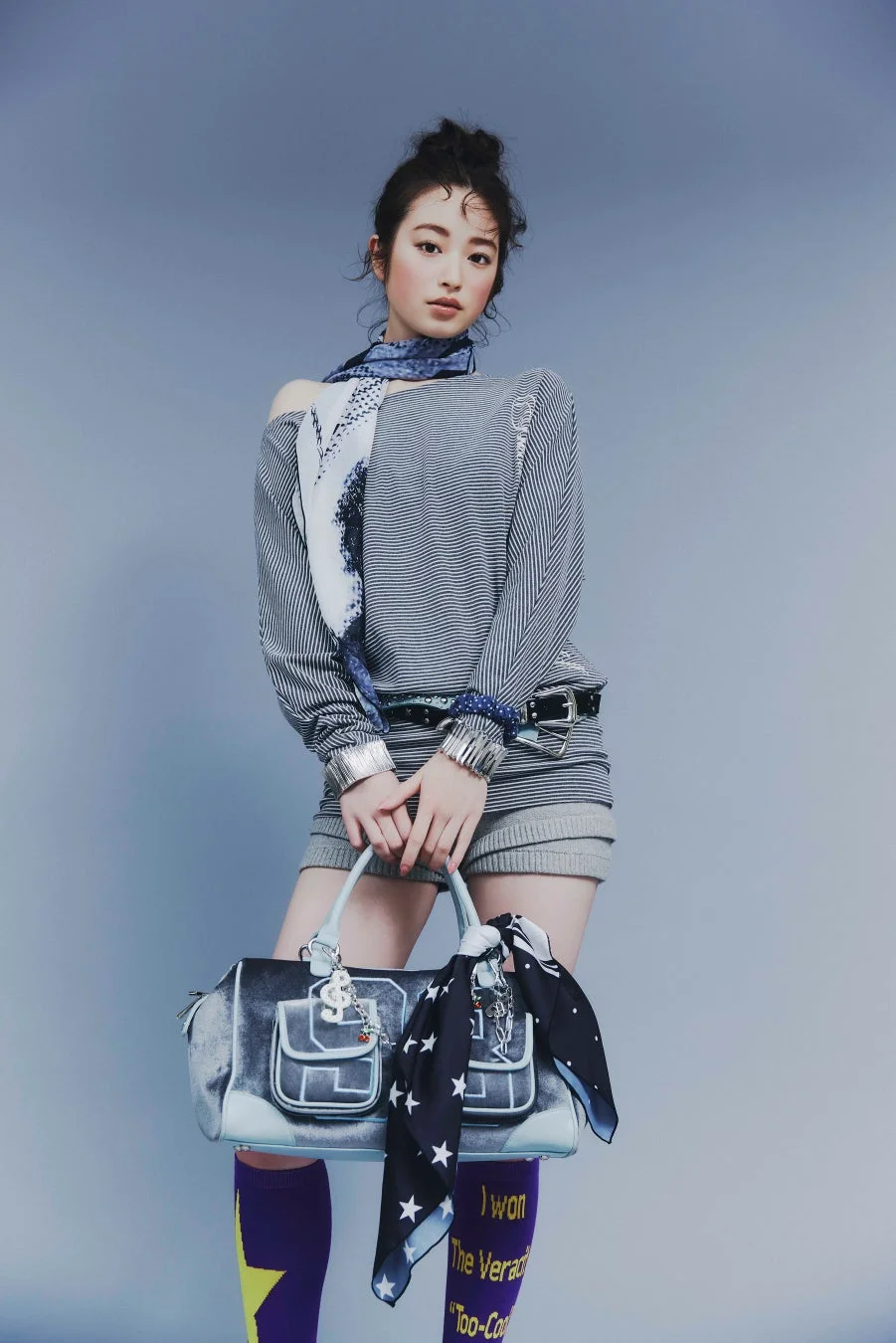 グレーの背景でポーズをとる若い女性のファッションポートレート。ストライプ柄のトップスとショートパンツを着用し、スカーフや個性的なロングソックス、大きなハンドバッグを合わせたスタイリッシュな装いです。