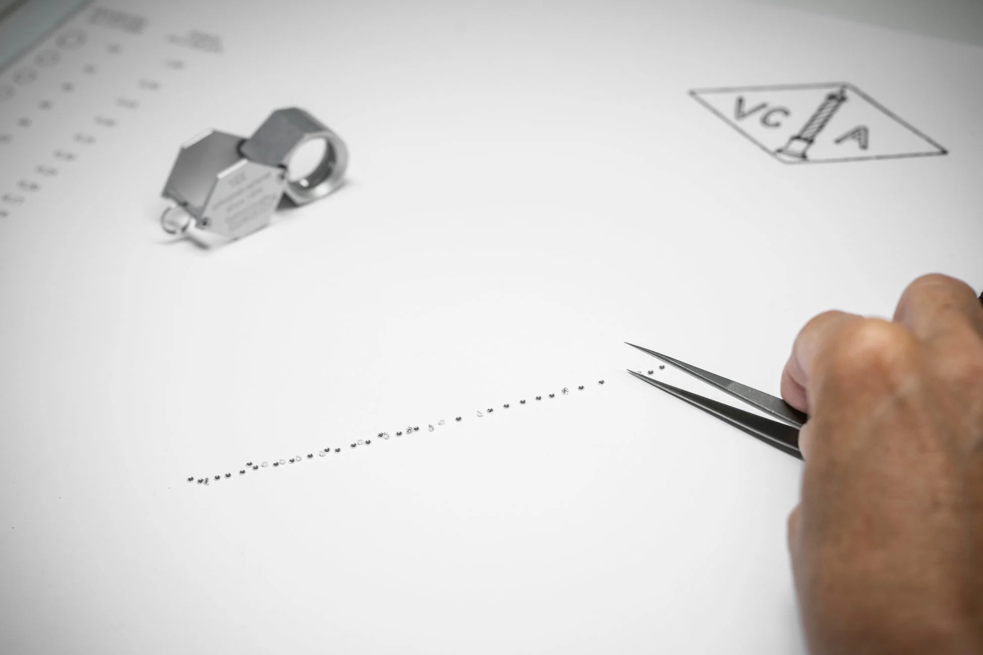 職人がピンセットを使い、白い台紙の上に小さなダイヤモンドや宝石を丁寧に並べている様子を捉えた画像。ルーペと「VC A」のロゴらしきスケッチも見られ、精密なジュエリー制作や選別作業の一端を示している。