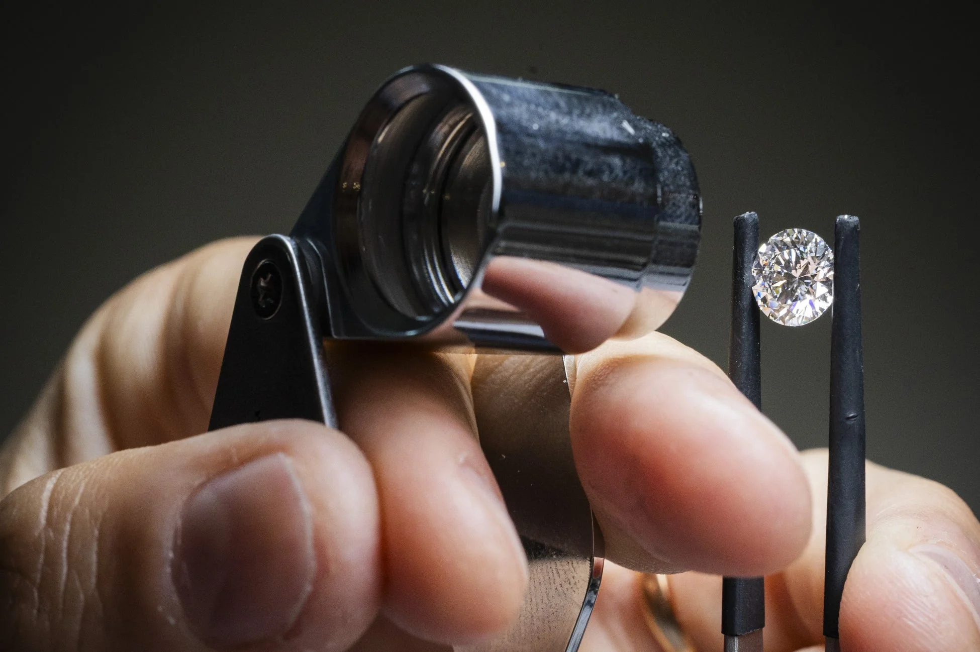 手がルーペとピンセットを使ってダイヤモンドを検査しているクローズアップ画像。宝石の品質鑑定や精査の様子を示しています。
