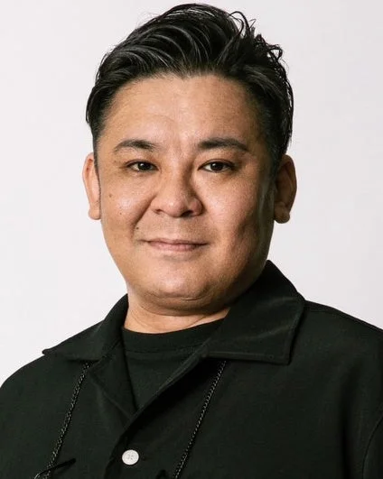 短い黒髪で黒いシャツを着たアジア人男性のポートレートです。男性はカメラをまっすぐ見ており、表情は穏やかで、背景はシンプルな白です。