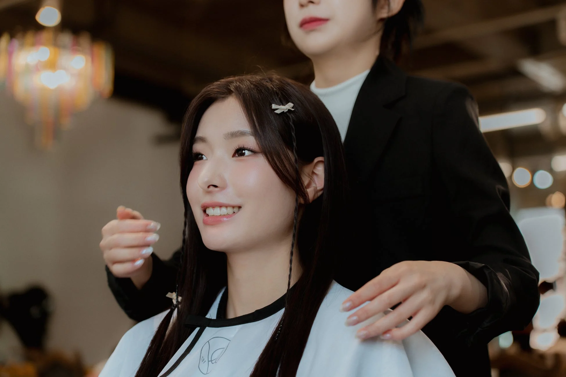 美容院で若い女性がヘアスタイリングを受けている様子。美容師が女性の髪に手を添え、女性は笑顔を見せている。ヘアクリップで髪が留められ、ヘアアレンジ中のようだ。