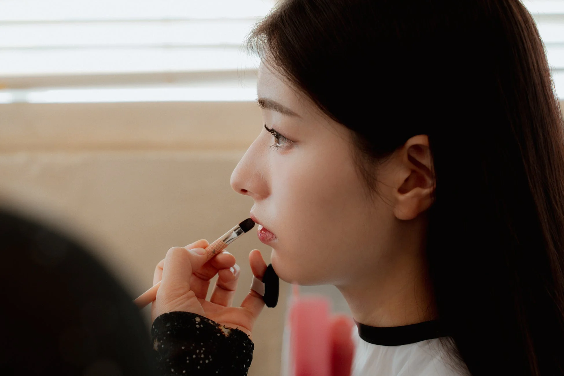 若い女性がメイクアップアーティストによってリップメイクを施されている横顔のクローズアップ写真。メイクブラシで丁寧に唇に色が塗られている瞬間を捉えています。