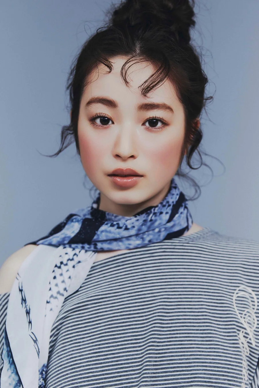 青い背景の前に立つ若い女性のポートレート。髪はまとめられ、ストライプのトップスと青いスカーフを着用し、穏やかな表情をしています。